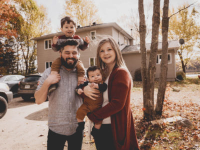 Matrimonio con hijos: padre y madre sonriendo delante de su vivienda unifamiliar mientras sostienen en brazos a sus dos hijos
