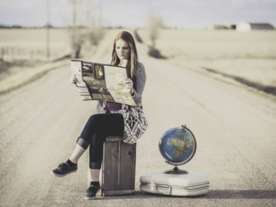 Mujer sentada sobre una maleta en medio de una carretera desierta consulta un mapa